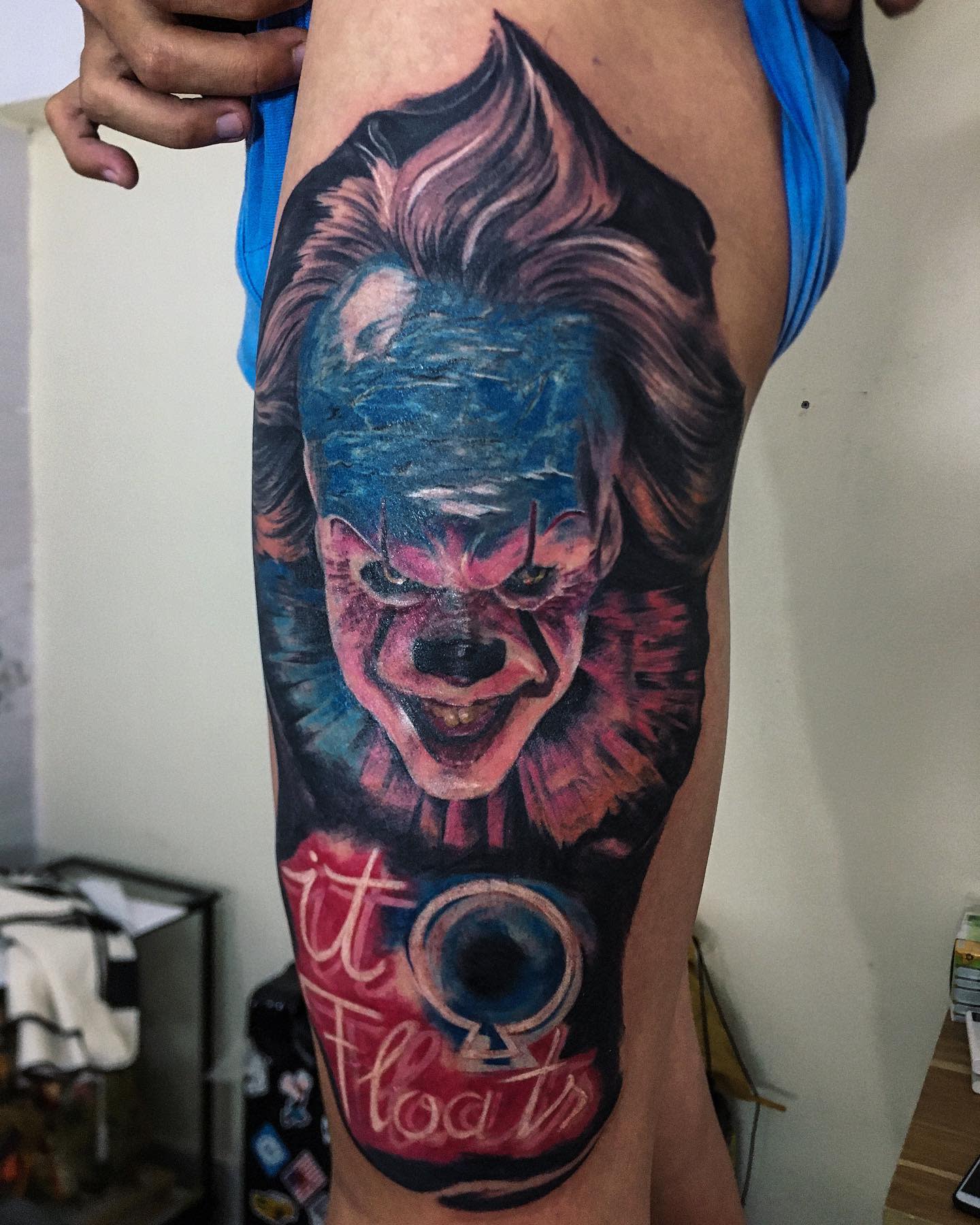 Colorful Pennywise tattoo on thigh with 'It Floats' text, featuring bold, vibrant hues